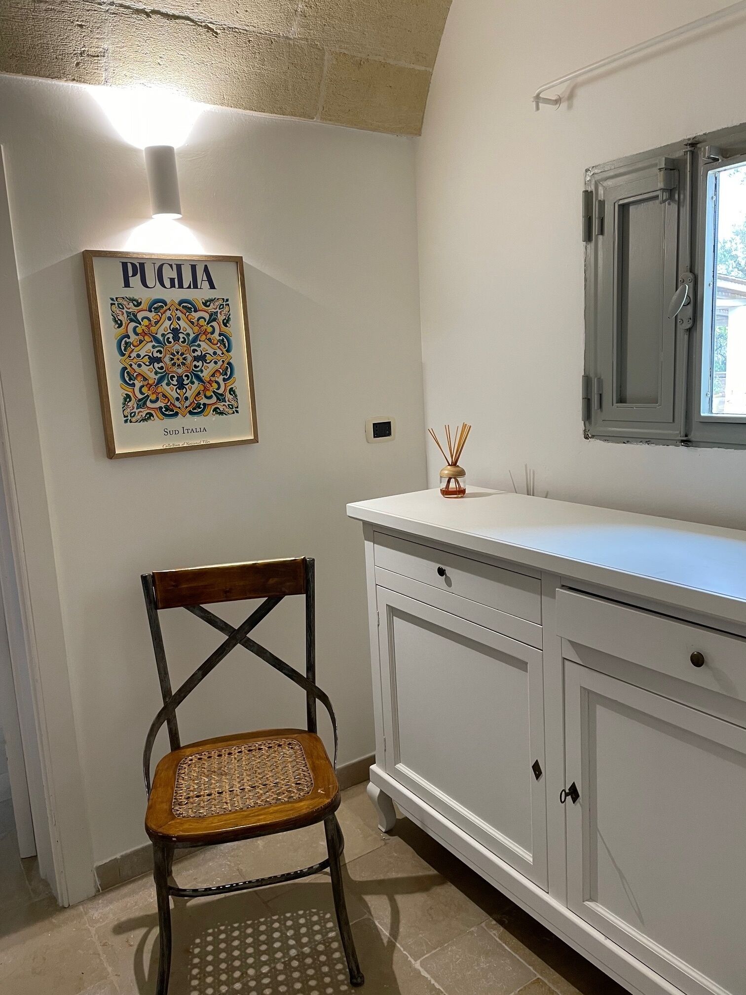 Una cocina con un armario blanco, una silla de madera, una obra enmarcada titulada 'PUGLIA Sud Italia' y una ventana con vistas a la vegetación.