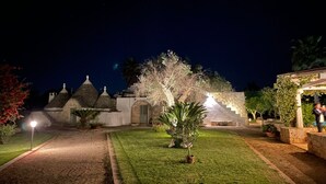 Vista nocturna de un patio con césped cuidado, edificios de piedra con techos cónicos y una pérgola llena de plantas.
