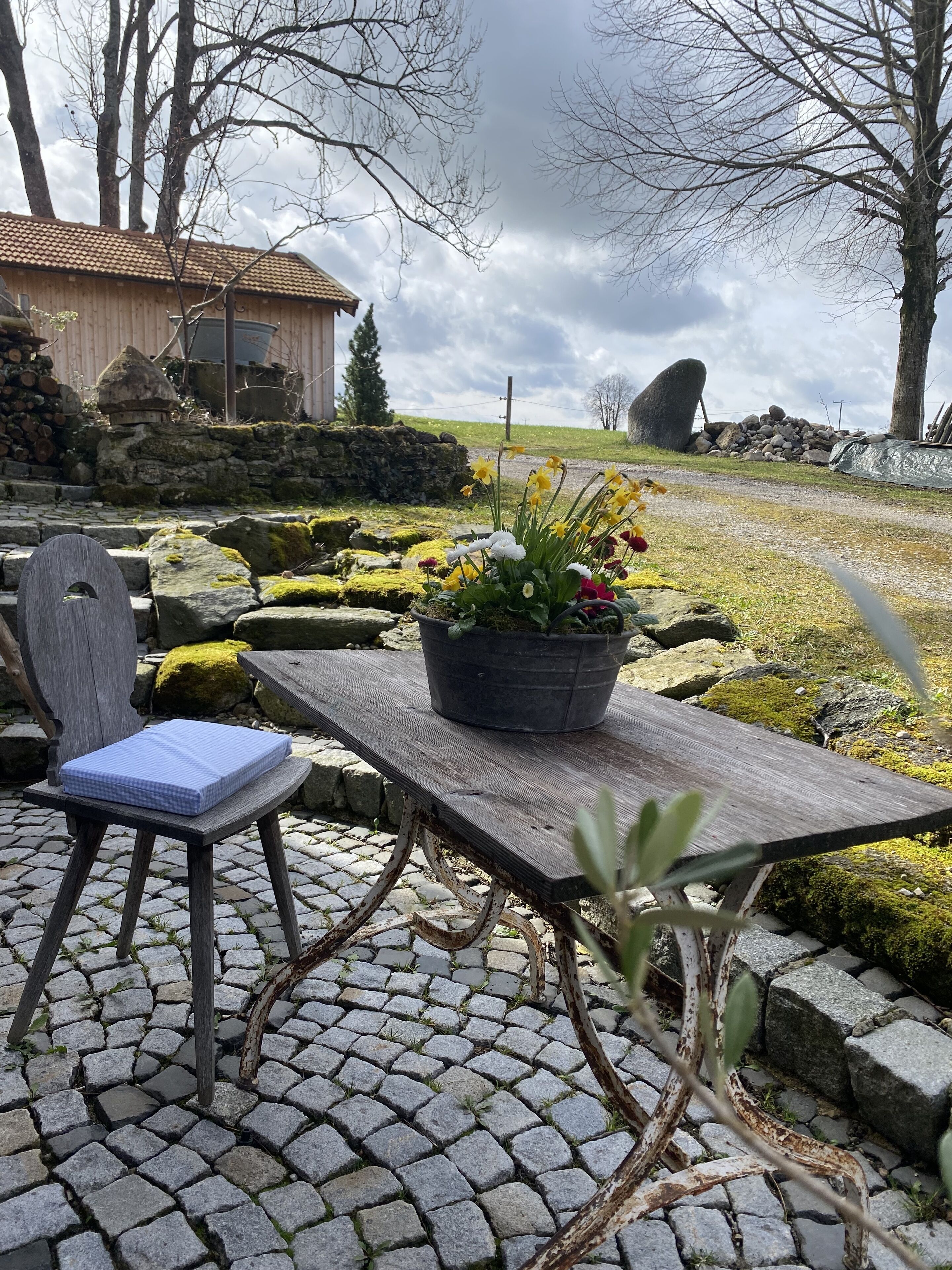 A rustic outdoor seating area with a wooden table and a chair, a potted plant, and a stone pathway.