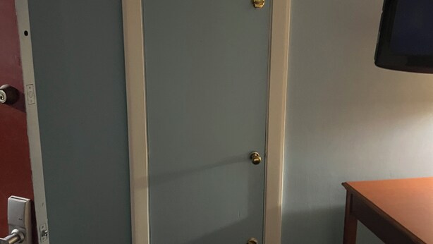 Ein Zimmer mit geschlossener Tür, einem kleinen Mülleimer, einem Holztisch und einem an der Wand montierten Fernseher.