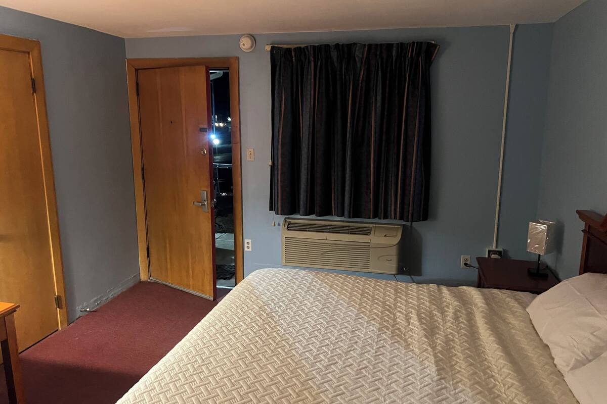 A hotel room with a bed, a window with curtains, an air conditioning unit, and a door.