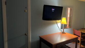 Habitació d'hotel amb una porta tancada, un televisor de pantalla plana fixat a la paret, una taula de fusta, una làmpada i una cadira.