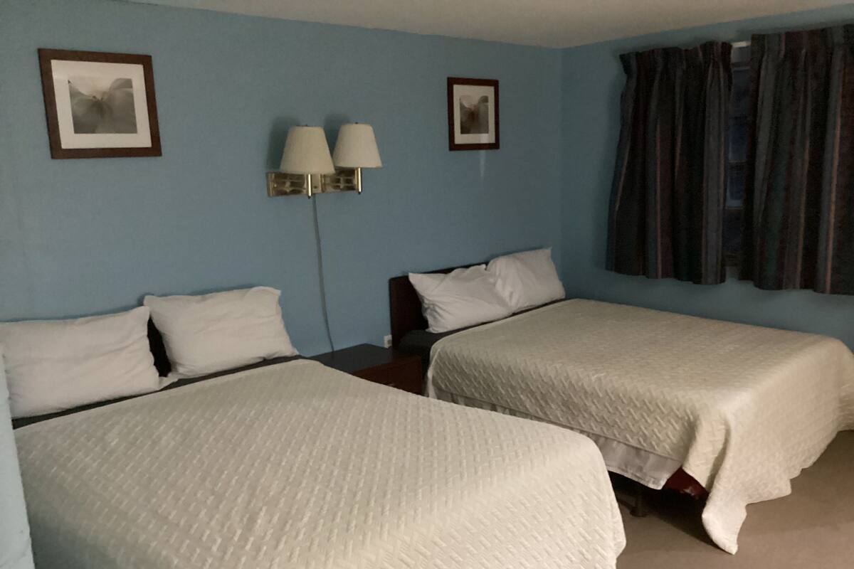 A hotel room with two beds, a nightstand, a lamp, and a window with curtains.