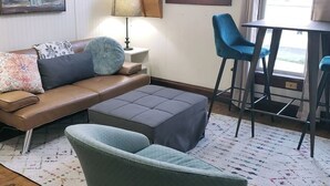 A living room with a brown leather sofa, a grey ottoman, a teal armchair, a small table with two blue chairs, a floor lamp, and a painting on the wall.