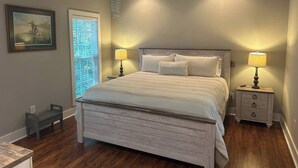 A bedroom with a large bed, two bedside tables with lamps, a small stool, and a window with blinds. - Refreshing & Peaceful Southern Inlet Beach House! (Murrels Inlet)