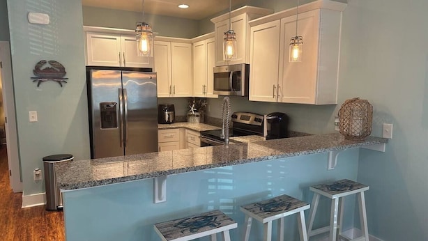 A modern kitchen with white cabinets, a stainless steel refrigerator, a microwave, and a granite countertop. There are three white barstools and pendant lights above the counter. - Refreshing & Peaceful Southern Inlet Beach House! (Murrels Inlet)