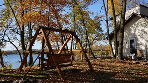 Un ensemble de balançoires en bois installé dans un espace boisé, avec une maison et un lac en arrière-plan.