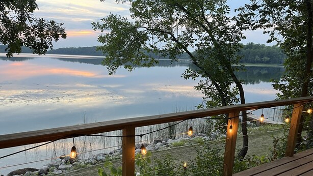 Une terrasse en bois dotée d’une balustrade, offrant une vue sur un lac paisible et une rive éloignée.
