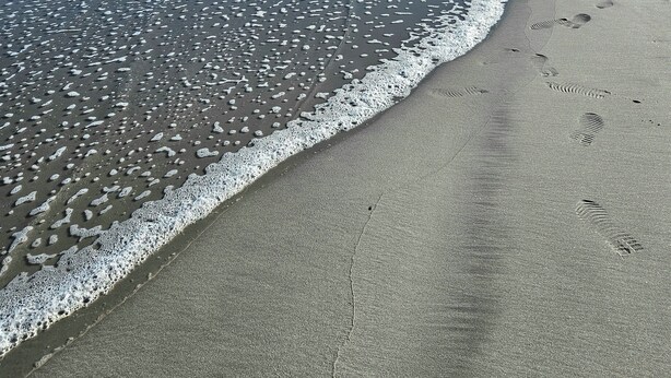 Uma praia com céu claro, ondas e pegadas na areia.