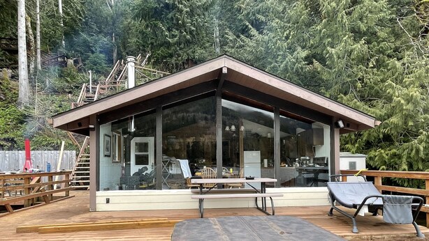A modern cabin with a glass front, wooden deck, and outdoor seating area. - Cozy Cabin on Lake Sutherland with hot tub, huge dock and kayaks! (Port Angeles)