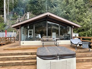 A modern cabin with a glass front, wooden deck, and outdoor seating area. - Cozy Cabin on Lake Sutherland with hot tub, huge dock and kayaks! (Port Angeles)