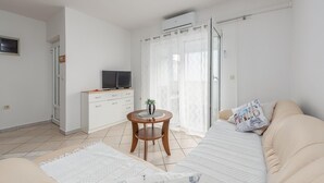 Una sala de estar moderna con un sofá color crema, una pequeña mesa de madera con una planta, un televisor sobre un mueble blanco y un ventanal con cortinas traslúcidas.