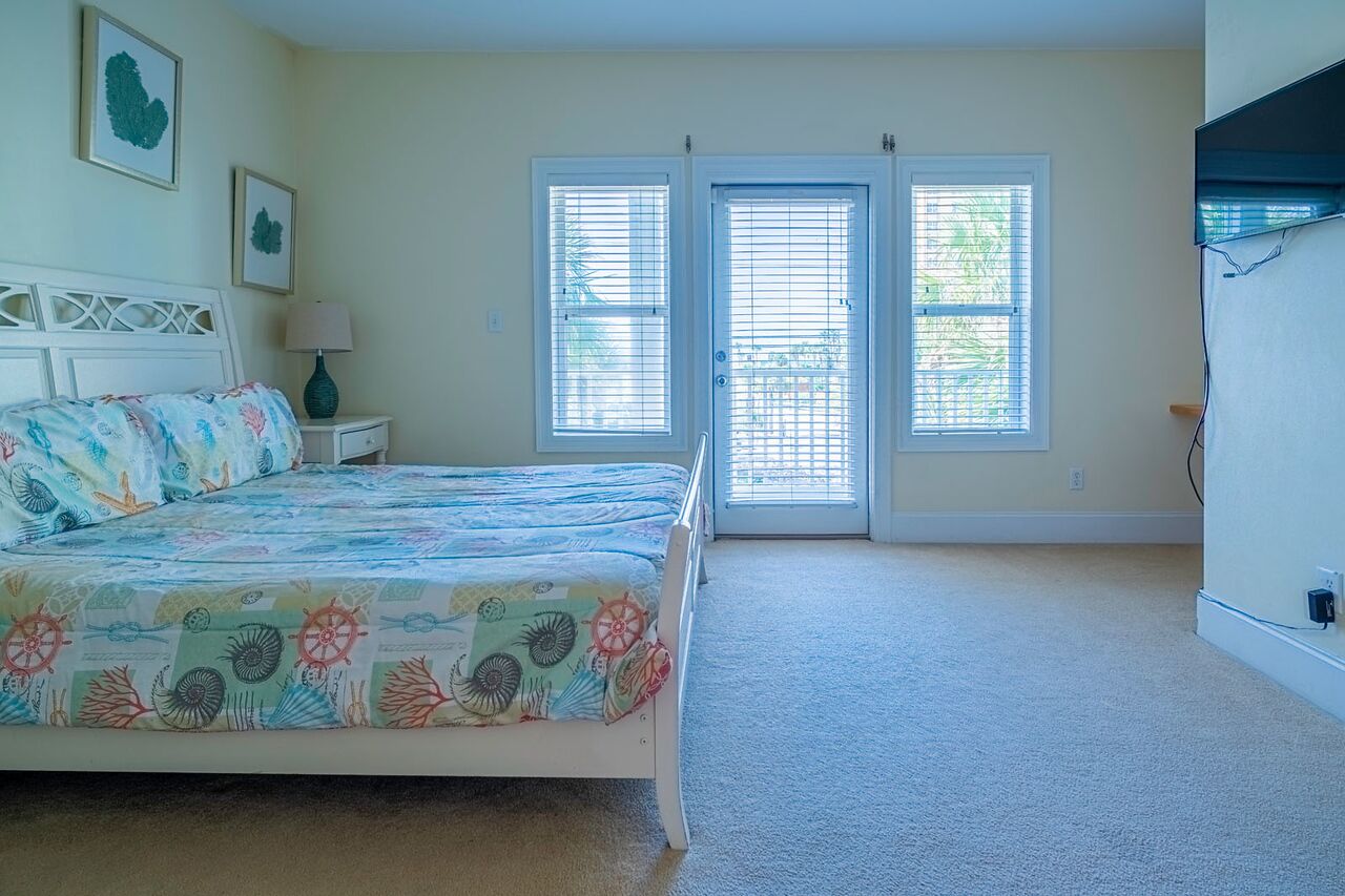 A bedroom with a bed, two windows with blinds, a nightstand with a lamp, and a wall-mounted TV.