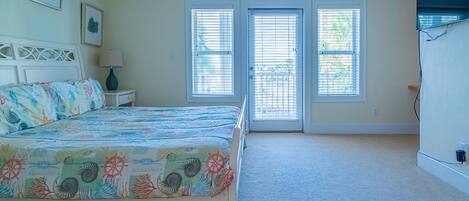 A bedroom with a bed, two windows with blinds, a nightstand with a lamp, and a wall-mounted TV.