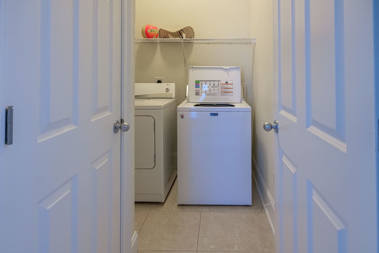 A laundry room with a washing machine and dryer, a shelf with a red ball, and a white refrigerator.
