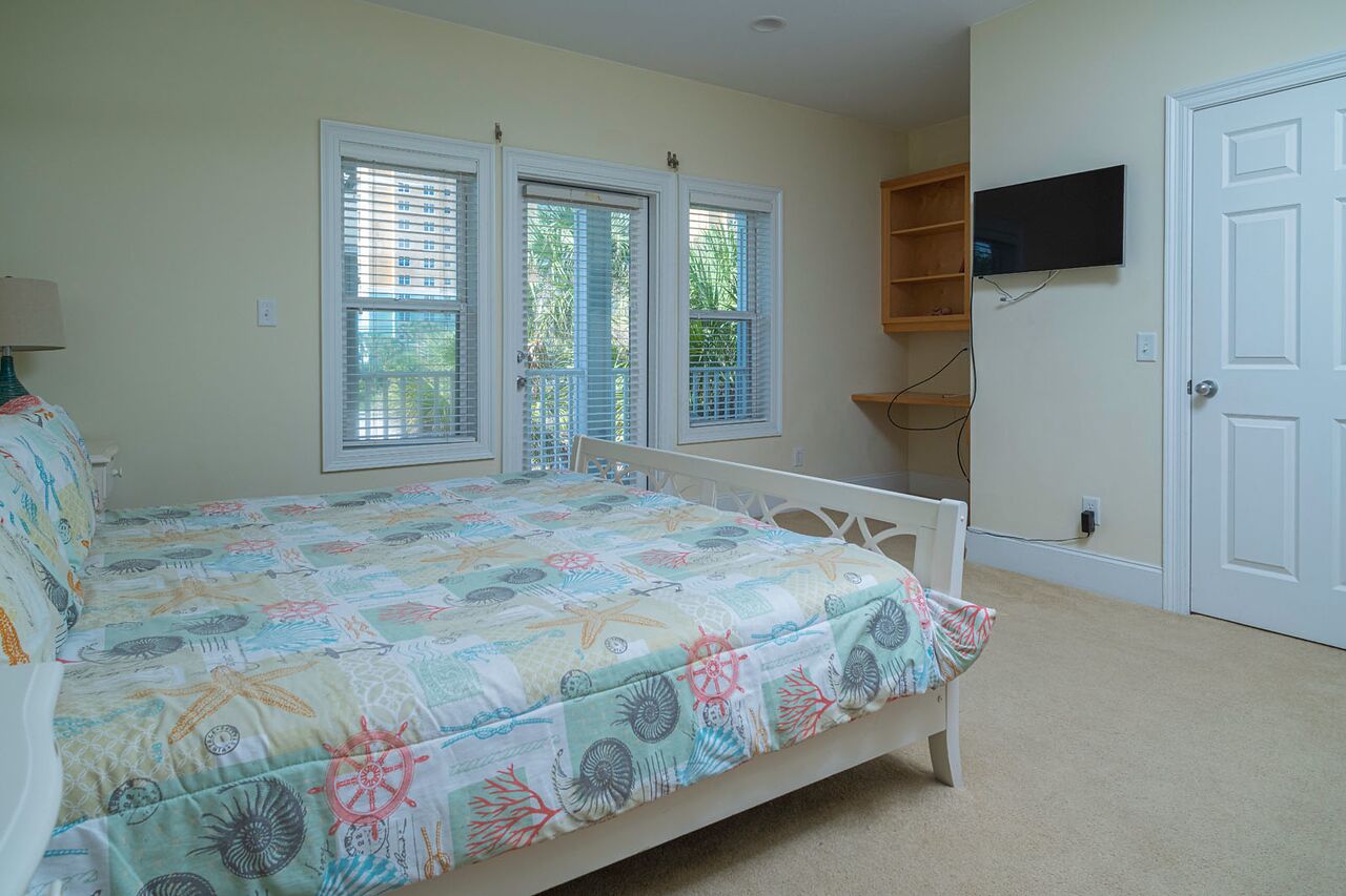 A bedroom with a bed, two windows with blinds, a TV on the wall, and a door.