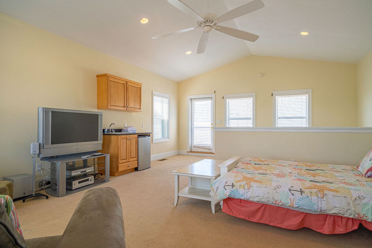 A bedroom with a bed, a TV, a ceiling fan, a small table, and a kitchenette.