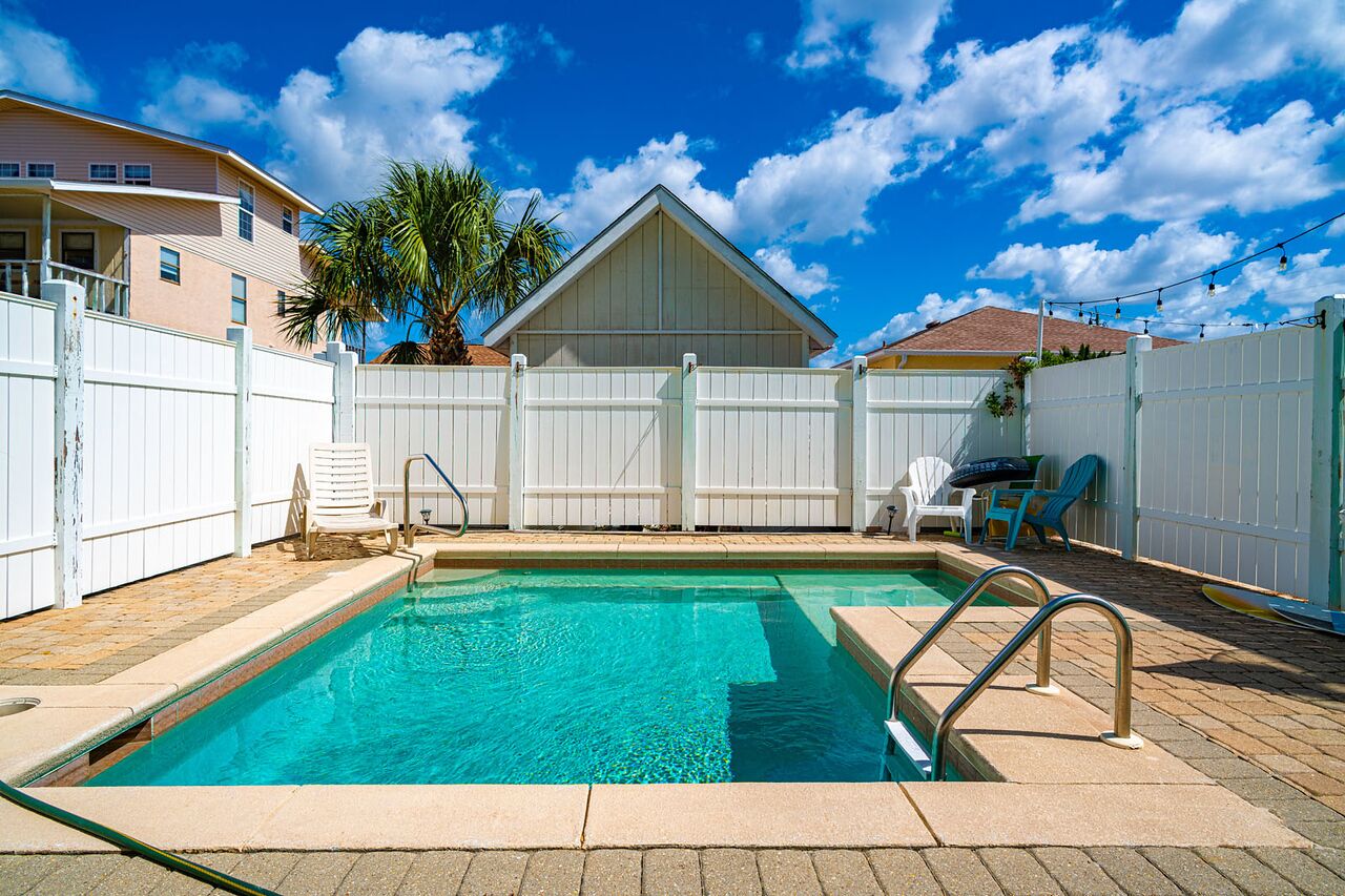A swimming pool with a white fence, a palm tree, and a house in the background.