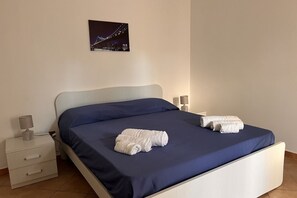 A neatly made bed with a blue bedspread and two rolled towels, a bedside lamp, and a small drawer unit.