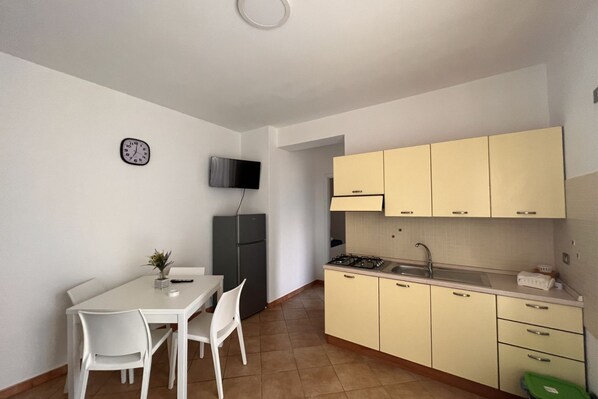 A compact kitchen with a dining table and chairs, a small refrigerator, and a TV mounted on the wall.