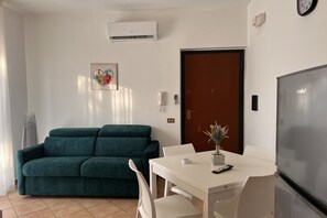 Ein Wohnzimmer mit einem türkisfarbenen Sofa, einem weißen Esstisch mit Stühlen, einem Fernseher und einer Tür mit einer Uhr.