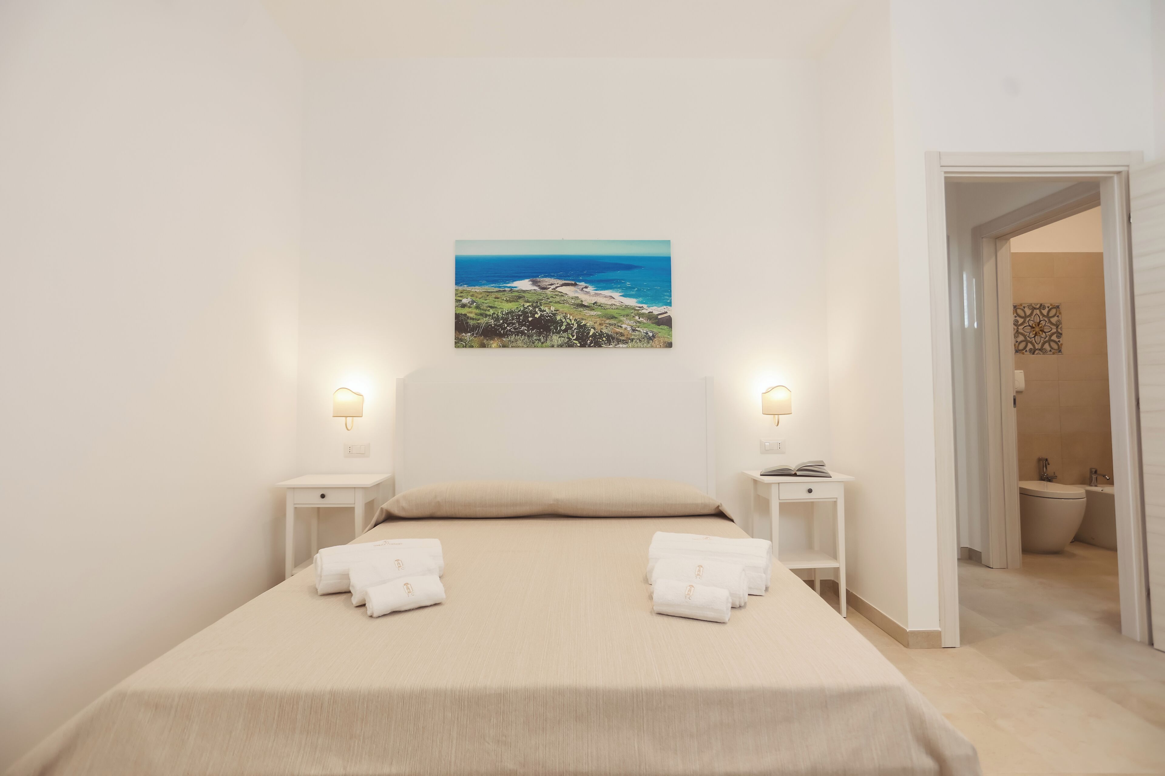 Una camera da letto con un letto, due comodini, un quadro che raffigura un paesaggio costiero e un bagno visibile attraverso una porta aperta.