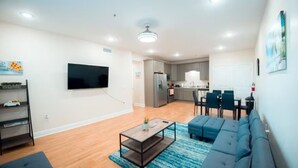 Ein modernes Wohnzimmer mit einer Couch, einem Couchtisch und einem Fernseher. Es gibt eine Küchenzeile mit Schränken und einem Kühlschrank.