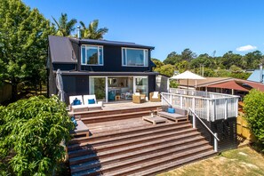 A two-story house with a deck, outdoor furniture, and a glass door entrance. - Newly renovated waterfront home, with large flat lawn leading to the beach. (Waiheke Island)