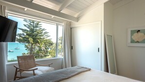 A bedroom with a bed, a chair, a TV, and a window with a view of the sea. - Newly renovated waterfront home, with large flat lawn leading to the beach. (Waiheke Island)