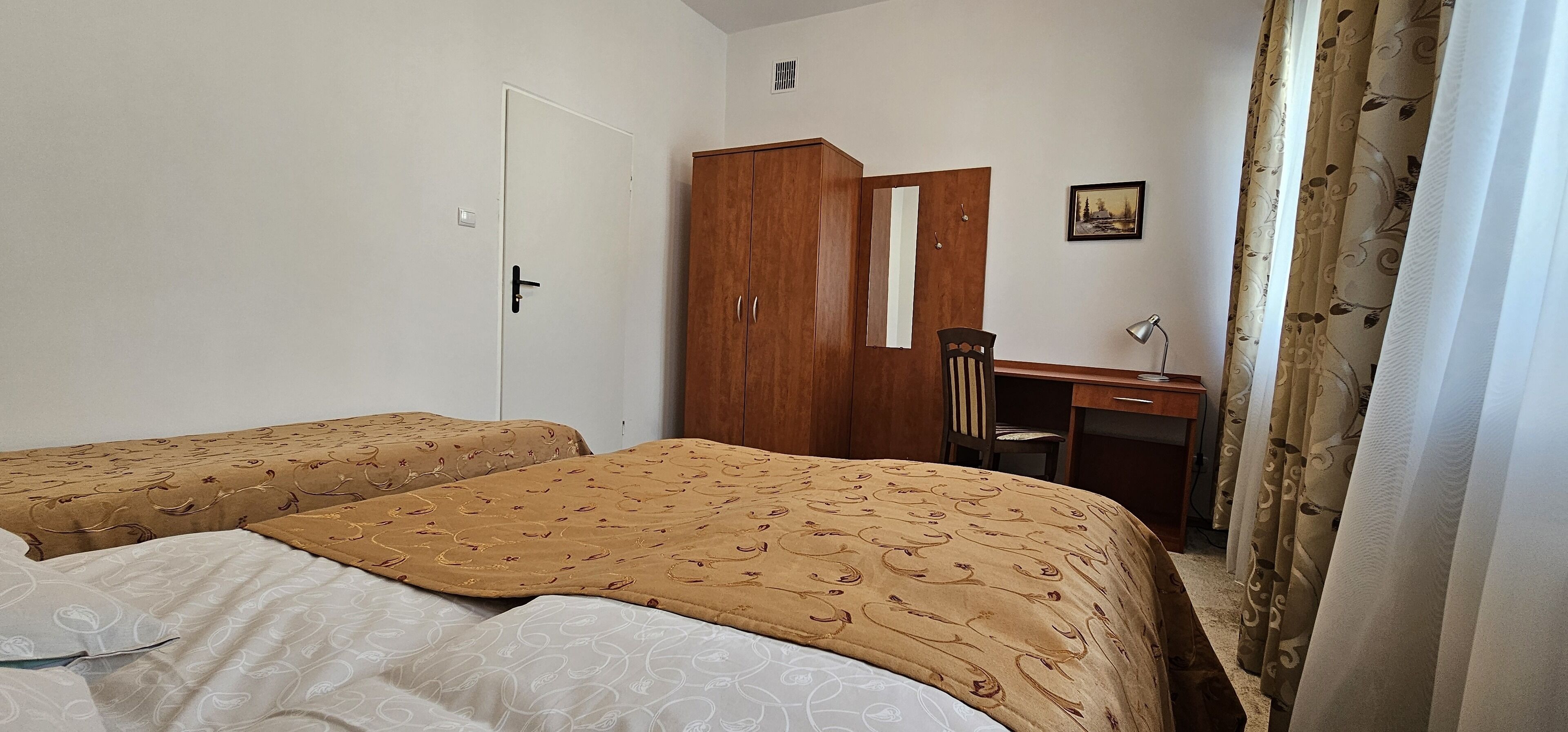 A bedroom with two beds, a wooden wardrobe, a desk with a chair, and a single window with curtains.