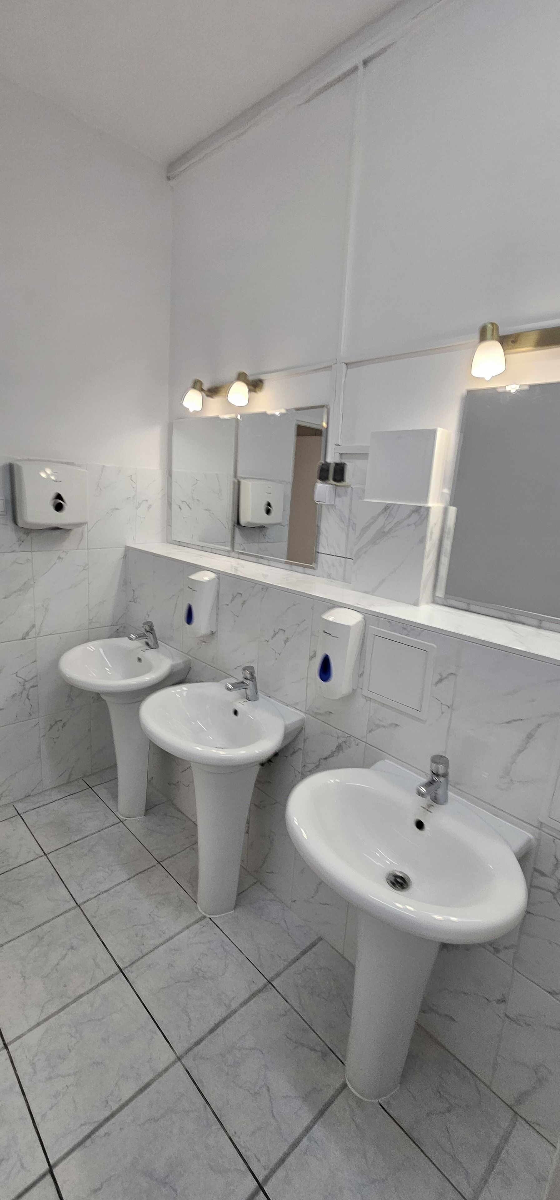 A bathroom with three sinks, marble-tiled walls, and wall-mounted soap dispensers.