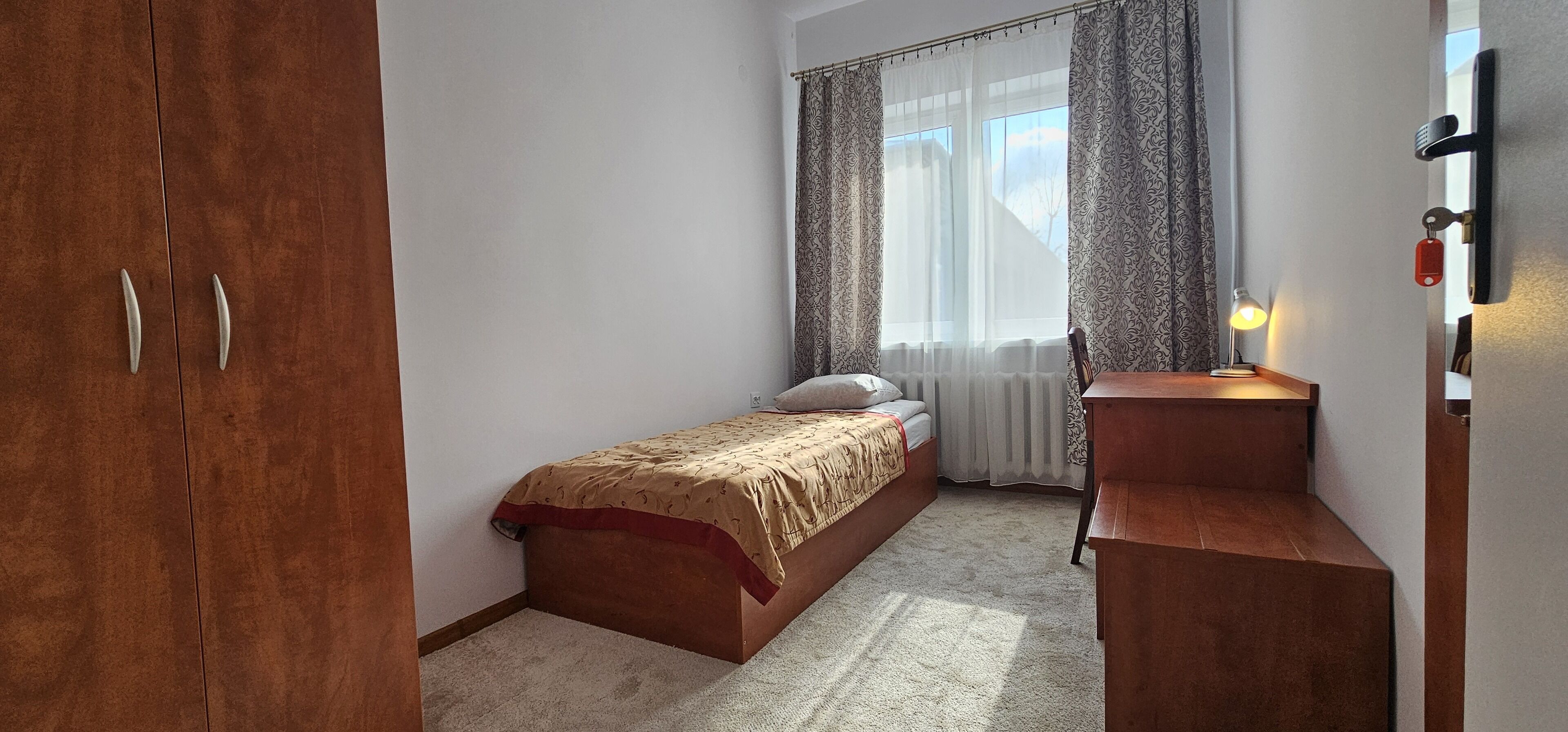 Un dormitorio con una cama individual, un armario de madera, una mesita de noche con lámpara y una ventana con cortinas.