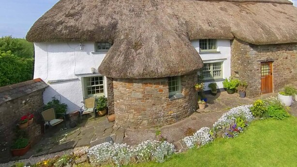 Una casa tradicional con techo de paja y una extensión de piedra, un jardín con plantas en macetas y una puerta de madera.