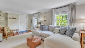 Un salon avec un canapé beige, une chaise en bois, un pouf marron et une porte blanche.