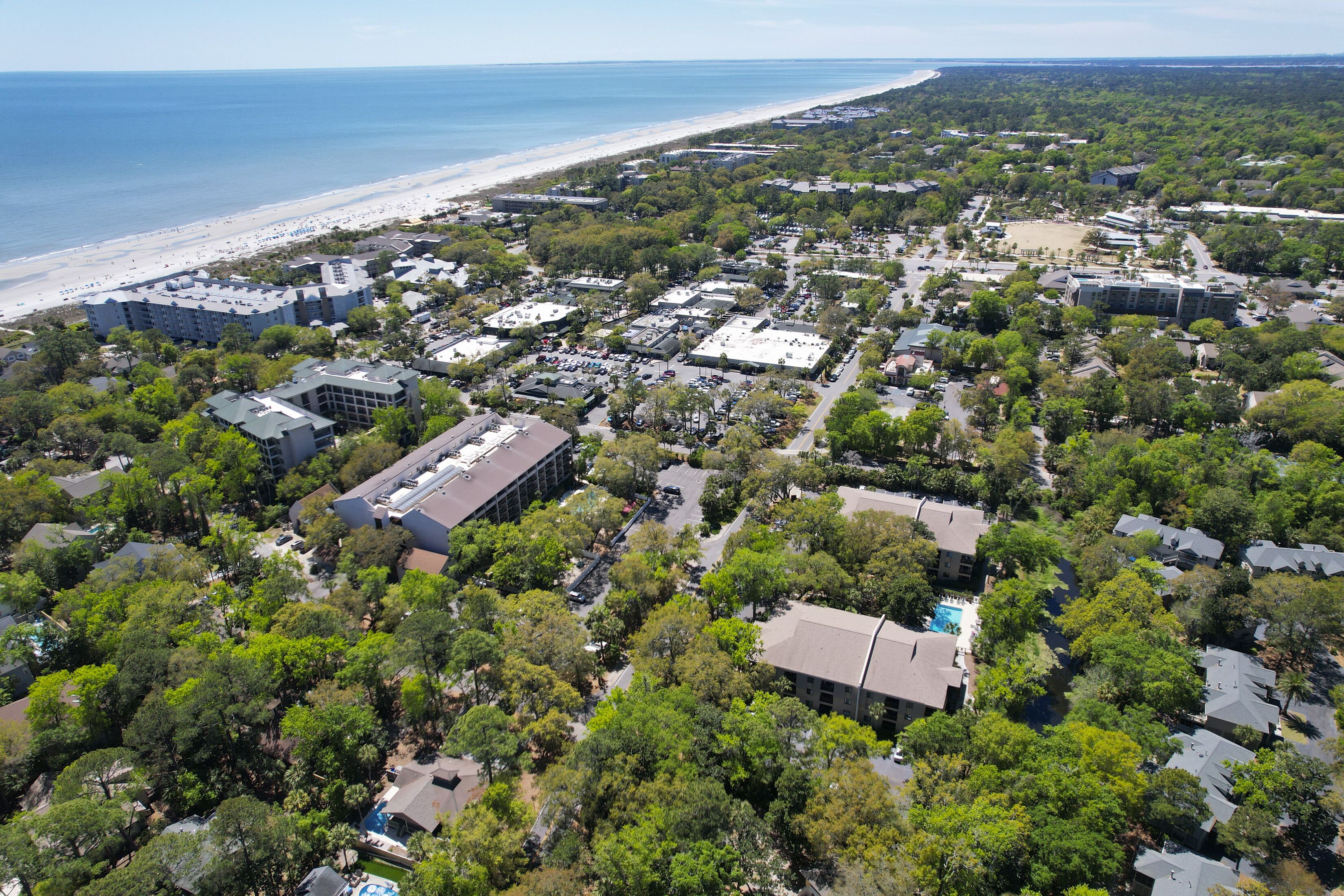 An aerial view of a coastal town with a beach, dense greenery, and various buildings including residential houses and commercial structures.
