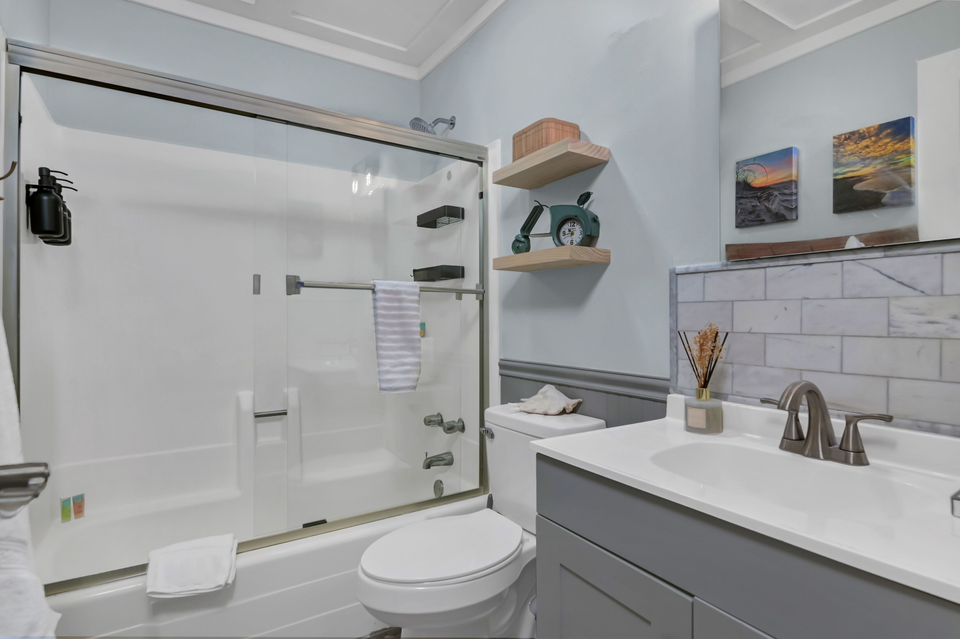 Een moderne badkamer met een douche in een glazen cabine, een wit toilet, een wastafel met een marmeren blad en decoratieve planken met klokken en kunstwerken.