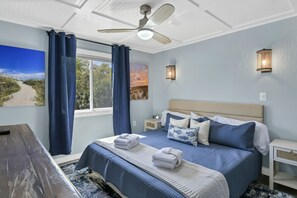 Un dormitorio con una cama grande, dos mesitas de noche, un ventilador de techo y una ventana con cortinas.