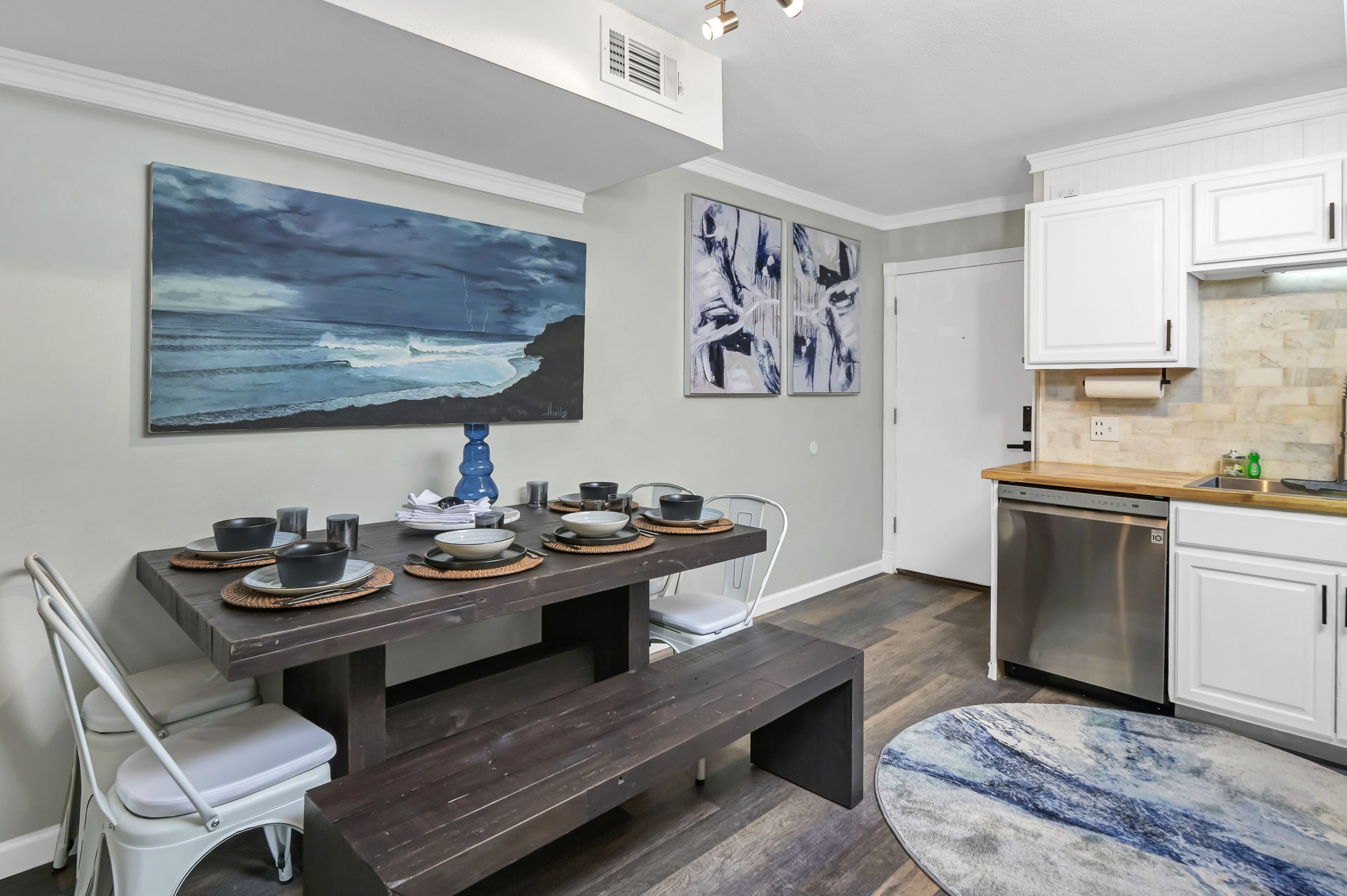 A modern kitchen with white cabinets, a stainless steel dishwasher, and a dining area with a dark wooden table set for four.