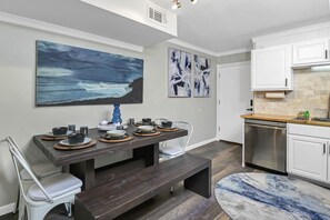 Una cocina moderna con armarios blancos, un lavavajillas de acero inoxidable y un comedor con una mesa de madera oscura dispuesta para cuatro comensales.