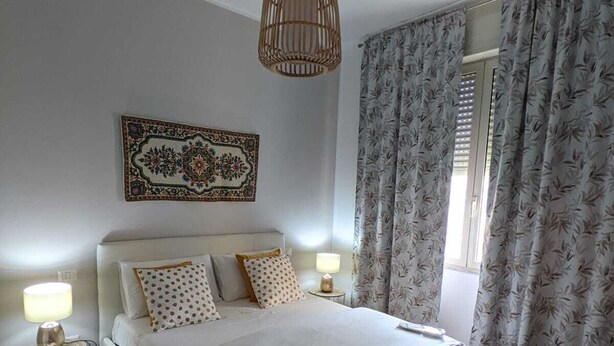 Un dormitorio ordenado con una cama grande, una lámpara colgante, un tapiz de pared con motivos y una ventana con cortinas.