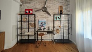 Una habitación con pared de ladrillo, dos estanterías metálicas negras con diversos objetos, un taburete de madera y un ventilador colgante.