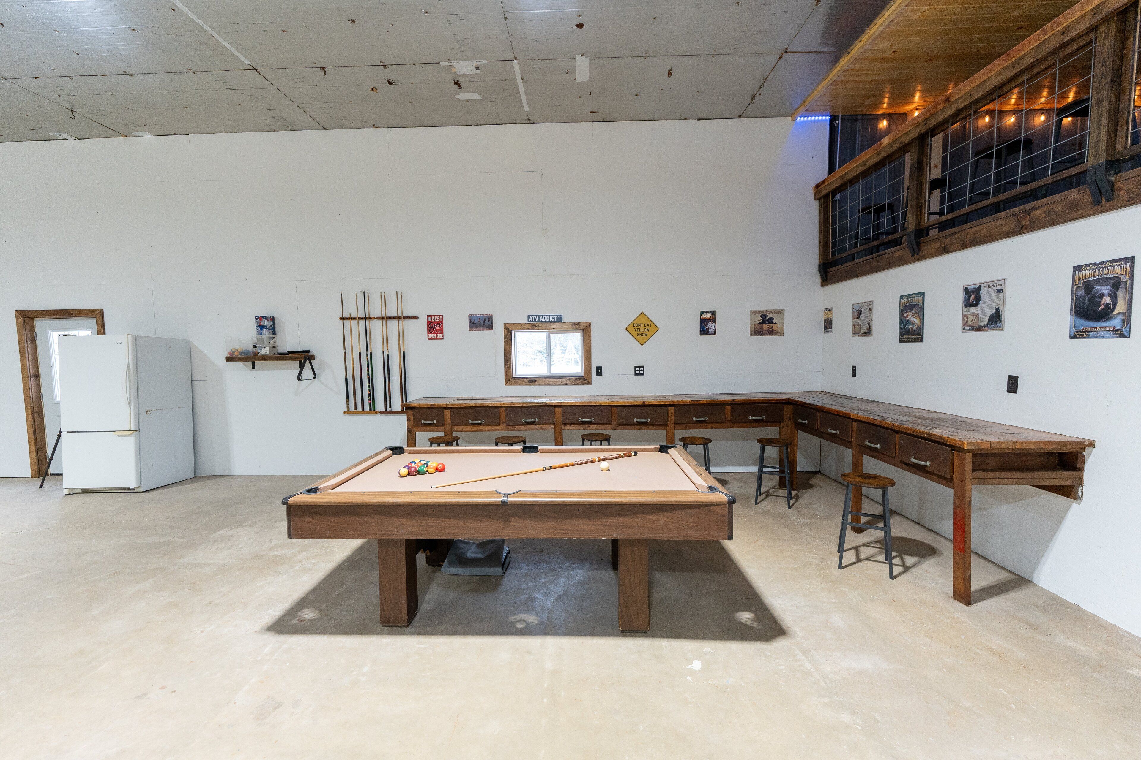 A pool table in a room with a wooden desk, a refrigerator, and a window.