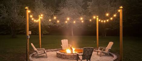 A circular fire pit with chairs around it, surrounded by a gravel area, with string lights above.