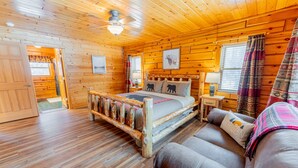 A wooden cabin bedroom with a bed, a wooden headboard, a nightstand, a lamp, and a plaid blanket on a couch.