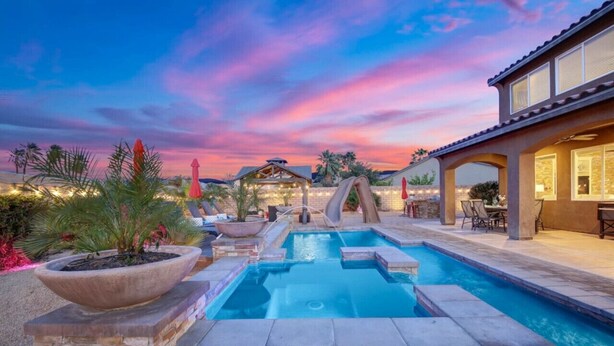 A swimming pool with a stone border, surrounded by potted plants and a patio area with outdoor furniture. - Carebnb/Waterslide/Casita/Game Room (Indio)