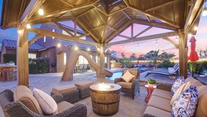 An outdoor patio area with wicker furniture, a fire pit, and a pool in the background. - Carebnb/Waterslide/Casita/Game Room (Indio)