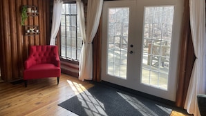 A cozy room with a red chair, wooden walls, a ceiling fan, and large windows with curtains. - The little Log Lodge (City of the Village of Clarkston)