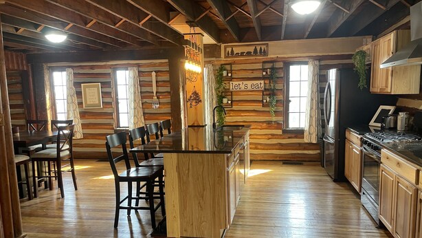 A rustic cabin interior with a wooden dining table and chairs, a kitchen with wooden cabinets and appliances, and a sign that reads "let's eat". - The little Log Lodge (City of the Village of Clarkston)