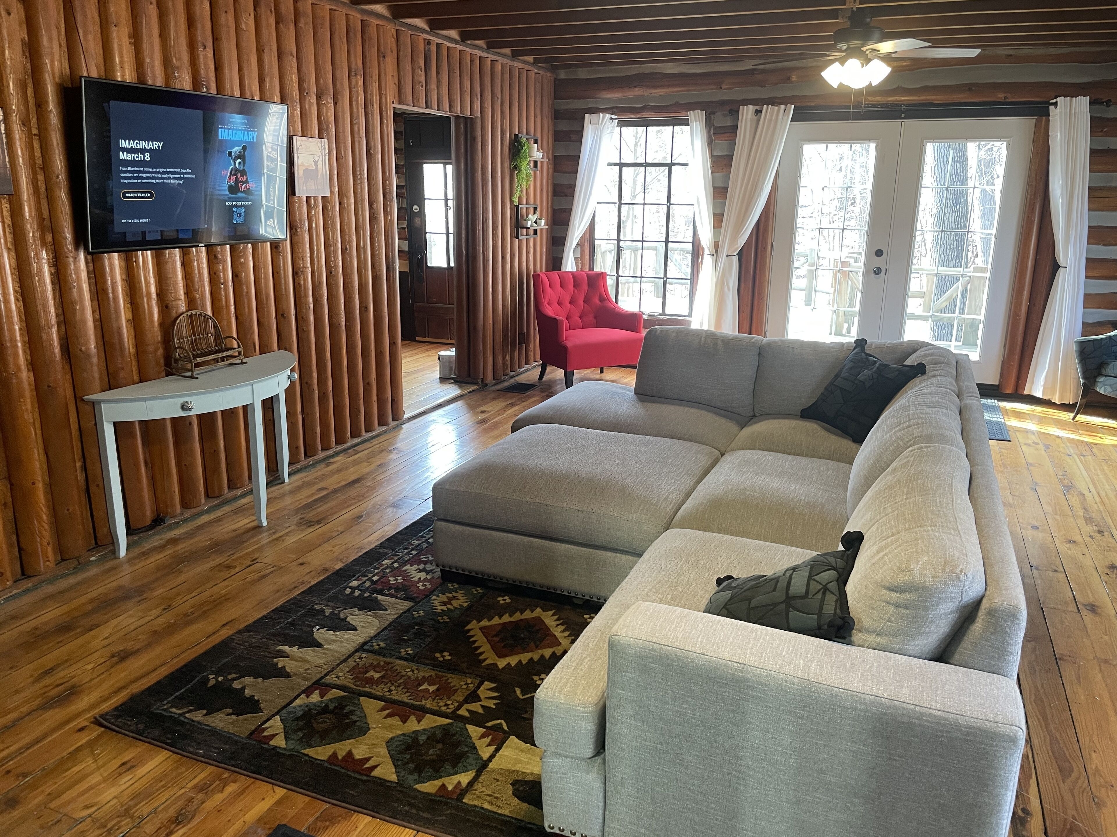 A cozy living room with a large sectional sofa, a red armchair, a small white table, and a flat-screen TV mounted on a wooden wall.