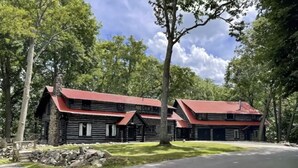 A log cabin with a red roof, surrounded by trees and a paved driveway. - The little Log Lodge (City of the Village of Clarkston)
